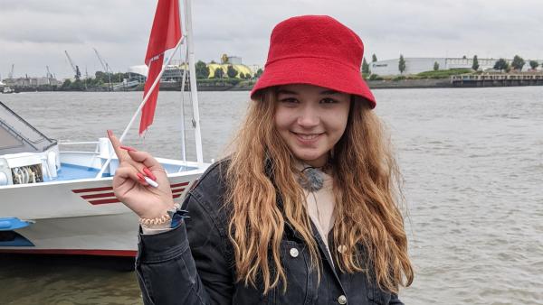 Rote Mütze Raphi steht mit einer roten Kopfbedeckung und roten Fingernägeln vor einem Fluss. Hinter ihr steht ein Boot. Sie lächelt.
