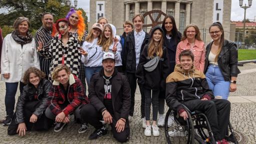 Jess ist beim Treffen der "Grünen Bande" dabei. Das Gruppenfoto zeigt die "Grüne Bande" und ist buntgemischt: verschiedene Haarfarben, Make-Up-Vorlieben und verschiedene Alter. Eine Person sitzt in einem Rollstuhl. Im Hintergrund sieht man die Volksbühne.