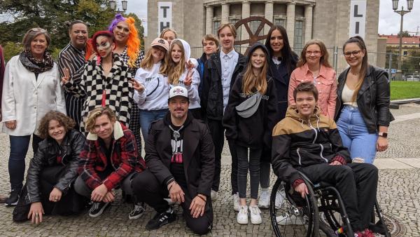 Jess ist beim Treffen der "Grünen Bande" dabei. Das Gruppenfoto zeigt die "Grüne Bande" und ist buntgemischt: verschiedene Haarfarben, Make-Up-Vorlieben und verschiedene Alter. Eine Person sitzt in einem Rollstuhl. Im Hintergrund sieht man die Volksbühne.