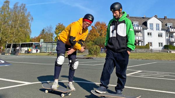 Ben, links, mit Helm, Schützern an Ellenbogen und Knien, steht mit einem Fuß auf einem Skateboard. Neben ihm steht ein Coach des Gorilla-Teams, ebenfalls mit Helm und einem Skateboard vor sich.