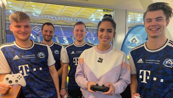 Im Hintergrund ist das HSV-Stadion zu sehen. Jess zwischen vier Männern in blauen HSV-Trikots, alle halten Controller in der Hand.