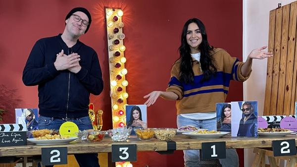 Ben (links) und Jess (rechts) stehen hinter einem Tisch. Auf dem Tisch liegen verschiedenes Essen und Bilder von Stars. Am Tisch hängen Nummern.