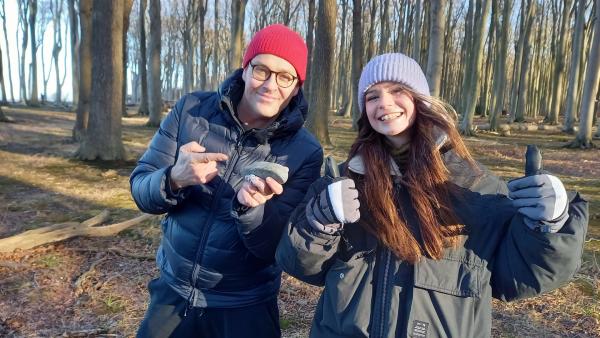 In einem Wald stehen Ben und Nadine Breaty. Ben hält einen Stein in der Hand und zeigt mit der anderen Hand darauf. Sarah lächelt in die Kamera und zeigt mit beiden Händen Daumen hoch.