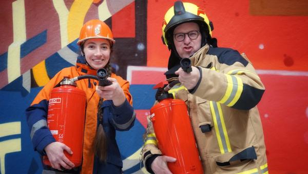 Ben und Emilia in Feuerwehr-Montur. Sie halten beide einen Feuerlöscher und richten die Öffnung in die Kamera.