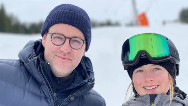 Ben steht rechts neben Snowboard-Freestyler Philippa. Philippa trägt einen Skihelm und eine Skibrille. Im Hintergrund ist eine Ski-Piste.