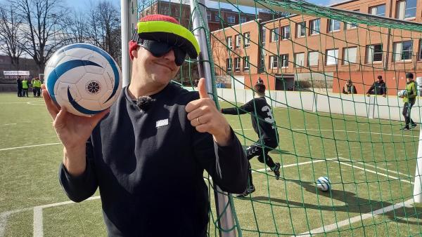 Ben steht hinter einem Tor auf einem Fußballplatz. In einer Hand hält einen Fußbal hoch. Mit der anderen Hand zeigt er einen Daumen hoch. Er trägt eine Brille mit verdunkelten Gläsern.