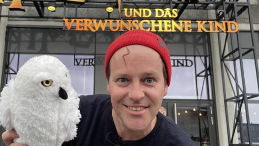 Ben steht vor dem Theater. Er hat ein Stofftier in der Hand und zeigt darauf. Es ist eine Eule aus den Harry Potter Filmen. Hinter ihm ist der Eingang des Theaters sichtbar. In großen Buchstaben ist "Harry Potter und das verwunschene Kind" zu lesen.