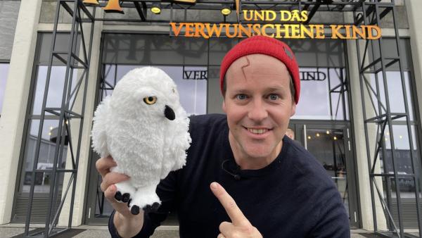 Ben steht vor dem Theater. Er hat ein Stofftier in der Hand und zeigt darauf. Es ist eine Eule aus den Harry Potter Filmen. Hinter ihm ist der Eingang des Theaters sichtbar. In großen Buchstaben ist "Harry Potter und das verwunschene Kind" zu lesen.