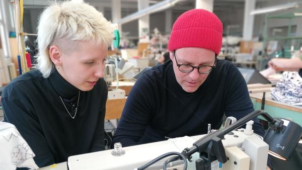 Im Vordergrund ist eine Nähmaschine. Dahinter sitzen Ben (links) und Sara Linke, die konzentriert einen Stoff vernähen.