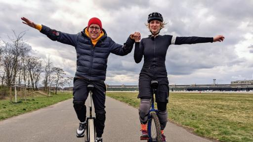 Auf einem Fahrradweg sind Ben (links) und Jana Tenambergen auf Einrädern unterwegs. Sie halten sich gegenseitig an den Händen und strecken den anderen, freien Arm aus.