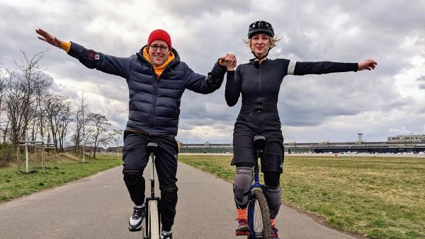 Auf einem Fahrradweg sind Ben (links) und Jana Tenambergen auf Einrädern unterwegs. Sie halten sich gegenseitig an den Händen und strecken den anderen, freien Arm aus.