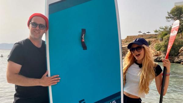 Ben trifft Mavie Noelle am Strand der Insel Mallorca. Mavie Noelle steht sichtlich begeistert mit Paddel in der Hand, Kappe auf dem Kopf und Blick in die Kamera, neben Ben. Ben hat ein Strandboard in der Hand und seine Mütze und Sonnebrille auf.