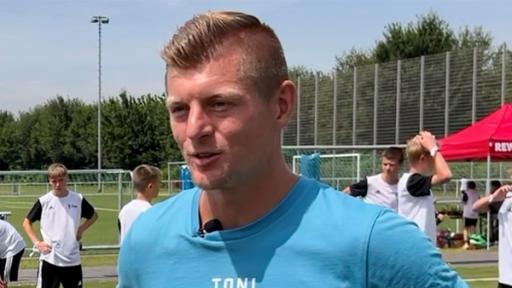 Links steht Sarah, die Toni Kroos ansieht, der rechts steht. Im Hintergrund sind Fußballplätze und jüngere Fußballspieler zu sehen.