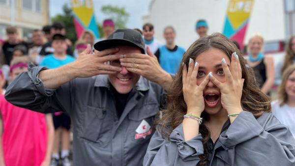 Vorne Sarah und Ben, die ihr Hände vor ihre Augen halten. Im Hintergrund sind Kinder zu sehen.