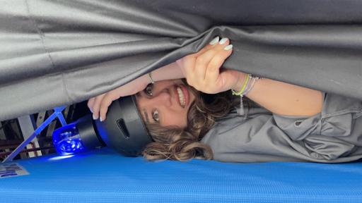 Auf einer blauen Matte liegt Sarah. Sie trägt einen Helm mit einem Blaulicht darauf. Mit ihrer Hand hält sie einen schwarzen von oben kommenden Vorhang hoch. Sie schaut verängstigt.