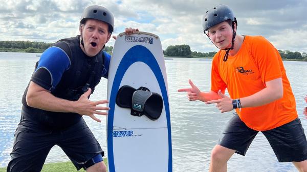 Links Ben, der einen Helm und ein Neoprenanzug trägt. Er stützt sich rechts auf ein Wakeboard. Rechts daneben steht Wakeboarder Fynn, der auf das Wakeboard in der Mitte zeigt.