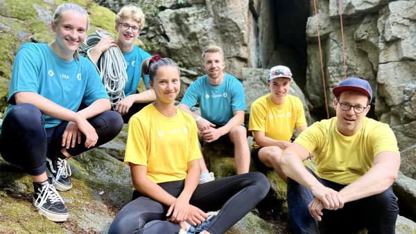 Alle stehen vor einer Felswand. Team "KiKA LIVE" in Gelb mit Alena, Felix und Ben und Team "Die Sportmacher" in Blau mit Lina, Otto und Stefan sind zu sehen. Otto hat ein noch gebundenes Kletterseil auf den Schultern.