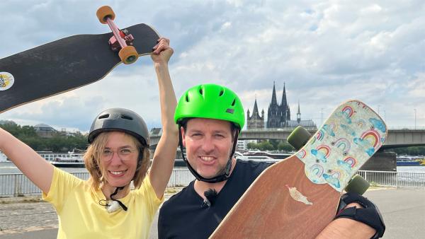 Deborah (links) und Ben stehen nebeneinander. Sie tragen Helme und halten jeweils ein Lonboard. Im Hintergund eine Brücke und ein Dom.