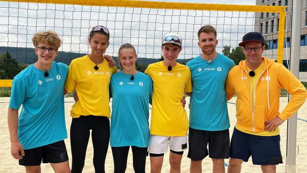 Die Teams beim Beachvolleyball. Von links nach rechts sind Otto, Alena, Lina, Felix, Stefan und Ben zu sehen. Sie stehen dicht beieinander und greifen sich um die Schultern und Rücken. Frontal fotografiert.