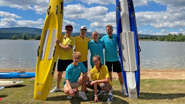 Beide Teams stehen vor dem Wasser, bereit für Runde zwei. Je zwei Personen aus jedem Team stehen im Hintergrund posierend. Im Vordergrund hockt je eine Person, damit alle auf dem Bild zu erkennen sind. Im Hintergrund werden die Boards festgehalten. 