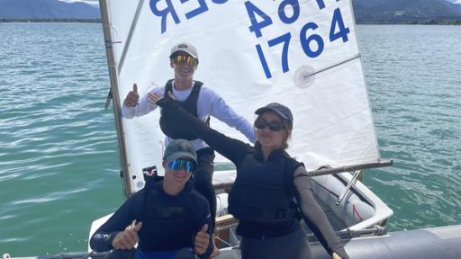 Auf einem  Segelboot auf einem Gewässer stehen Sarah (rechts) und die Zwillingsbrüder Lucas und Moritz (links). Sarah streckt die Arme aus. Lucas und Moritz zeigen einen Daumen nach oben.