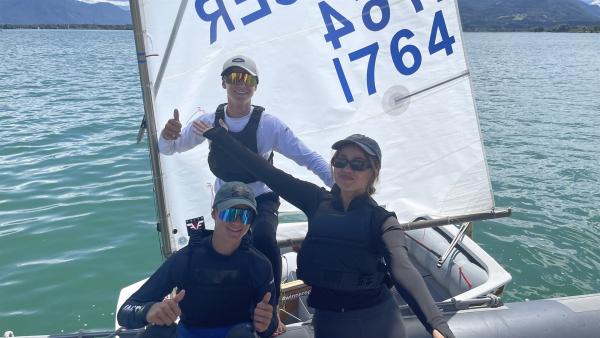 Auf einem  Segelboot auf einem Gewässer stehen Sarah (rechts) und die Zwillingsbrüder Lucas und Moritz (links). Sarah streckt die Arme aus. Lucas und Moritz zeigen einen Daumen nach oben.