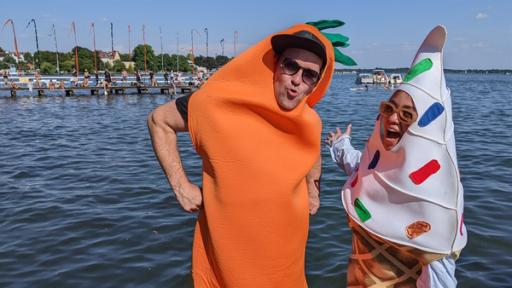 Ben und Sarah tragen Kostüme und stehen vor dem Müggelsse auf einem Steg. Ben ist als Möhre und Sarah als Softeis mit Streuseln verkleidet.