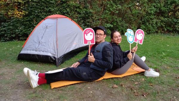 Im Grünen sitzen Ben (links) und Sarah (rechts) Rücken-an-Rücken auf einer orangenen Isomatte. Sie halten Holzstäbe mit aufgeklebten Emojis eines Mädchens und eines Jungen in ihren Händen. Im Hintergrund steht ein Zelt.