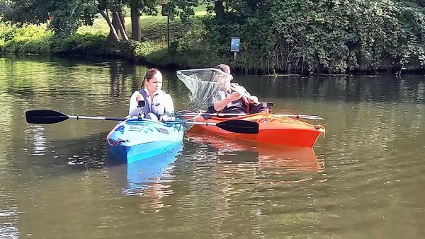 Sarah (links) und Ben sitzen auf einem Fluss jeweils in einem Kajak.
