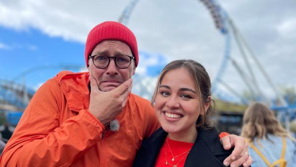 Ben und Sarah stehen nebeneinander. Sarah lächelt, während Ben das Gesicht verzieht und ein Hand am Kinn hält. Im Hintergrund ist ein Freizeitpark.