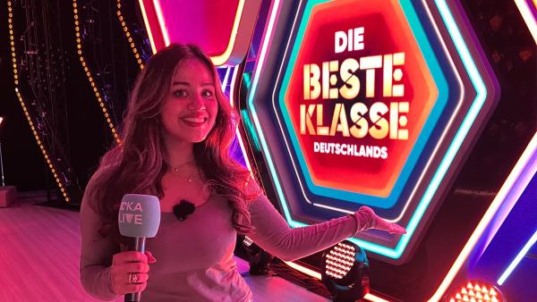 Sarah mit einem 'KiKA LIVE' Mikrofon steht in pink getauchtem Licht vor einem leuchtenden Schild mit der Aufschrift "Die Beste Klasse Deutschlands".