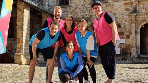  Von links stehen Sherif, Tim, Laura, Sarah, Annika und Ben in einem Innenhof eines Schlosses. Sherif, Sarah und Annika tragen blaue Leibchen während Tim, Laura und Ben pinke Leibchen tragen.