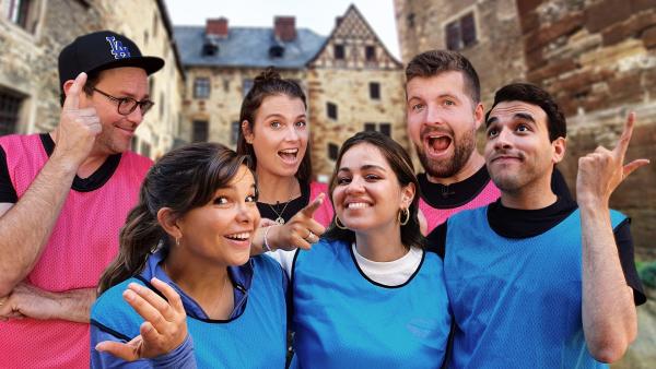 Vorne steht von links nach rechts Team blau Annika, Sarah und Sherif. Dahinter steht Team pink von links nach recht Ben, Laura und Tim. Im Hintergrund ist ein Schloss zu sehen.