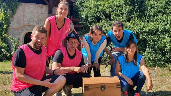 In der Mitte steht eine Holzkiste, um die Team blau und pink stehen. Von links steht in Team pink Tim, Laura und Ben. Von rechts steht Team blau Annika, Sherif und Sarah.