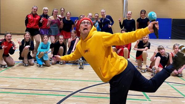 In einer Sporthalle steht Ben im Vordergrund, der eine einbeinige Pose macht und dabei eine Grimasse zieht. Hinter ihm steht und sitzt eine Gruppe Kinder, die Rollschuhe anhaben.