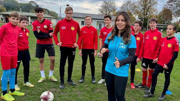 Jess steht in der Mitte mit einem blauen T-Shirt und hat eine Pfeife umhängen. Um sie herum stehen mehrere Jungs mit roten Jacken. Vor ihnen liegt ein Fußball. Im Hintergrund Rasen.