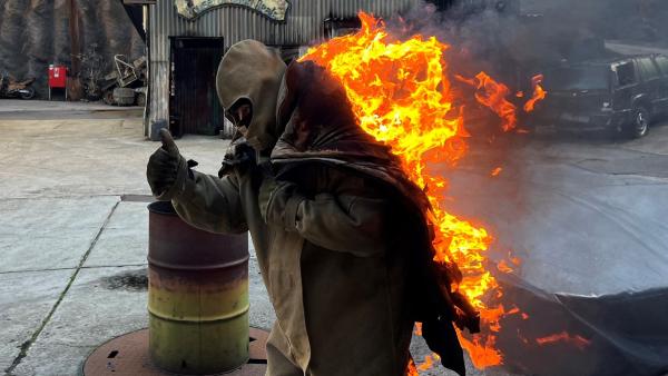 An einer Filmkulisse steht eine Person aus dem Stuntteam in Flammen. Sie signalisiert das alles okay ist. Die Person ist im Fokus des Bildes und ganz zentral abgebildet. Das Feuer ist am Rücken großflächig zu sehen. Die Person trägt Schutzkleidung, samt Kopfschutz.