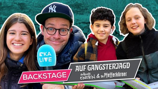 Ben steht als zweiter von links und hält ein KiKA LIVE Mikrofon. Links neben ihm steht ein Mädchen. Rechts von ihm stehen ein Junge und ein Mädchen. Vorne steht auf einem pinken Banner "Backstage" und auf einem grauen Banner daneben "Auf Gangsterjagd: Schloss Einstein und die Pfefferkörner".