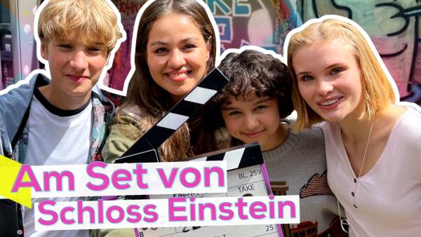 Moderatorin Sarah zusammen mit den Schloss Einstein-Darstellern Jonah, Janissa und Caroline. Vor ihnen eine Filmklappe und der pinke Schriftzug Am Set von Schloss Einstein. Sie lächeln in die Kamera. Hinter ihnen eine Wand mit Graffitti.
