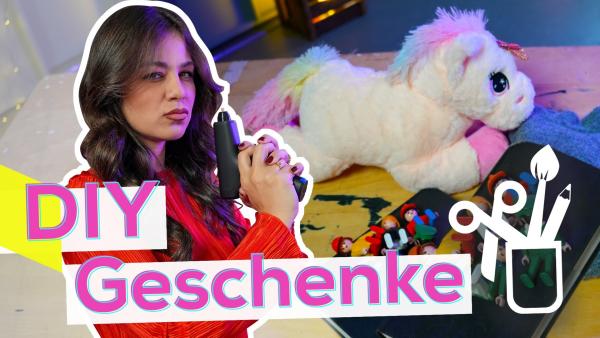 Links steht Sarah seitlich und hält eine Heißklebepistole vor sich. Im Hintergrund ein Tisch mit einem Plüsch-Einhorn, Stoff, kleinen Figuren und Büchern. Unten die Aufschrift DIY Geschenke, daneben eine weiße Grafik von Bastelutensilien in einem Behälter.