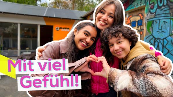 Sarah lächelnd in der Mitte. Sie umarmt links Sophia und rechts Janisa. Sophia und Janisa formen mit ihren Händen gemeinsam ein Herz. Aufschrift unten links Mit viel Gefühl!