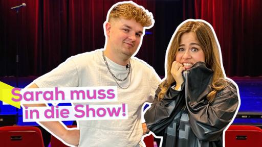 Comedian Tobii und Moderatorin Sarah stehen nebeneinander. Sarah mit ängstlichem Blick. Im Hintergrund die Bühne für den Auftritt. Aufschrift links: Sarah muss in die Show!