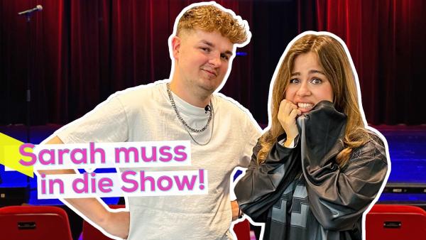 Comedian Tobii und Moderatorin Sarah stehen nebeneinander. Sarah mit ängstlichem Blick. Im Hintergrund die Bühne für den Auftritt. Aufschrift links: Sarah muss in die Show!