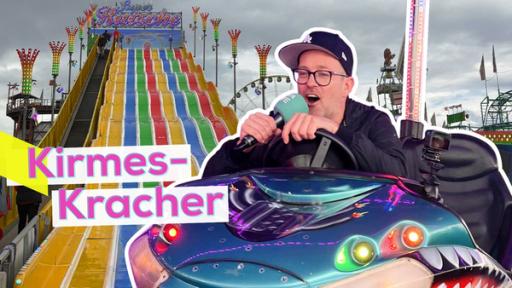 Rechte: KiKA/Rebekka Barta/Sabine Krätzschmar Ben, sehr fröhlich in einem Auto-Scooter. Im Hintergrund die Riesen-Rutsche auf dem Stuttgarter Frühlingsfest. Aufschrift: Kirmes-Kracher