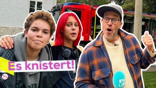 Ava und Elly eng nebeneinander, daneben Ben, der erstaunt mit dem Daumen hinter sich zeigt. Im Hintergrund ein Feuerwehrauto. Aufschrift: Es knistert!
