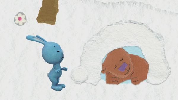Kikaninchen steht vor einer verschneiten Höhle, in der ein Bär schläft.