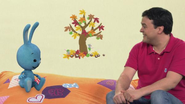 Kikaninchen und Christian reden über einen ein bunten Baum, im Hintergrund.
