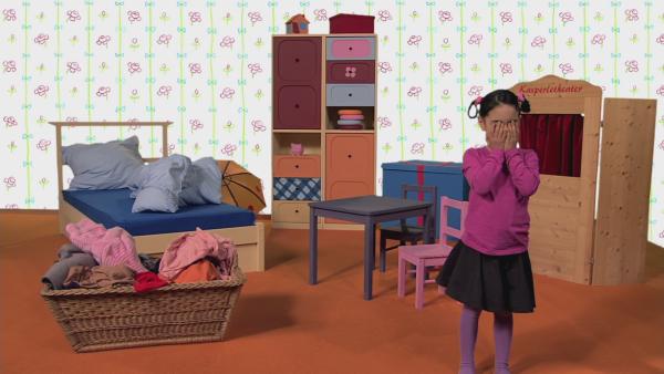 Manou steht im Kinderzimmer und hält sich die Augen zu. 