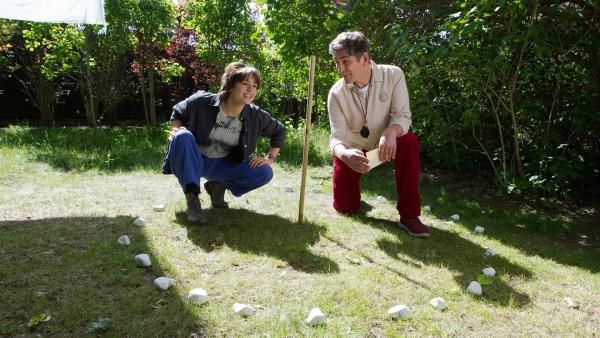 Fritz Fuchs und Elli knien auf dem Rasen in einer Sonnenuhr. Elli schaut auf den langen Schatten, den der Holzstab wirft und lächelt. Fritz Fuchs hält ein Stück Papier in den Händen und schaut zu Elli.