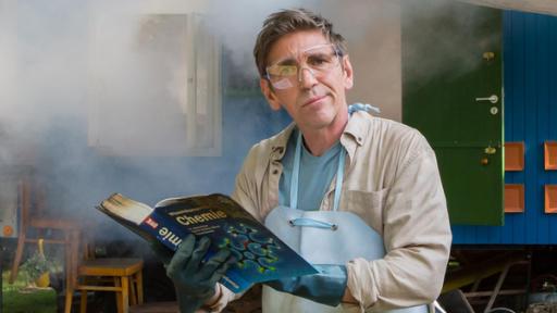 Fritz steht mit einer Schutzmaske und einem Chemiebuch vor dem Bauwagen, der in Rauch eingehüllt ist